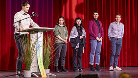 Five people are standing on a stage in front of a red curtain; one person is speaking at a lectern with a microphone, whilst the other four are standing side by side, looking straight ahead