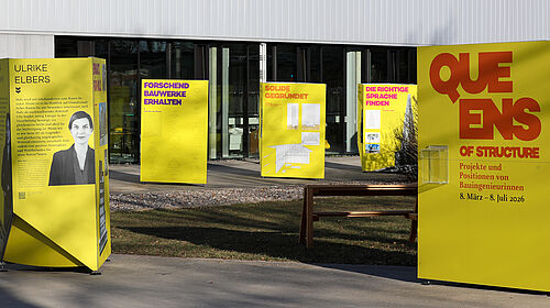 Es sind fünf rund zwei Meter hohe und ein Meter breite gelbe Rechtecke zu sehen. Auf diesen befinden sich Porträts von Frauen und TExt.