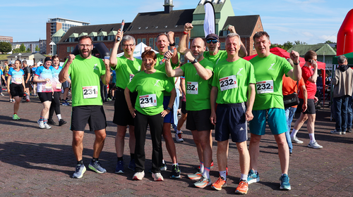 Acht Teilnehmende der Hochschule Wismar stehen in grünen Laufshirts beim Rostocker Firmenlauf 2025 zusammen, posieren gut gelaunt und mit erhobenen Armen für ein Gruppenfoto auf dem Veranstaltungsplatz. Sie tragen Startnummern mit den Teamnummern 231 und 232. Im Hintergrund weitere Läuferinnen und Läufer, Zelte und die Dächer der Haedgehalbinsel in Rostock.