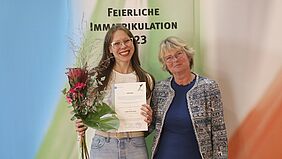 Die Preisträgerin ist gemeinsam mit der Leiterin des Internationalen Büros vor einer Bannerwand zu sehen, auf der "Feierliche Immatrikulation 2023" steht. Sie hält einen Blumenstrauß und die Urkunde in der Hand.