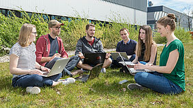 Im Bild sind sechs Studierende der Wirtschaftsinformatik zu sehen, die mit ihren Laptops auf der Campuswiese vor dem Haus 6 sitzen.