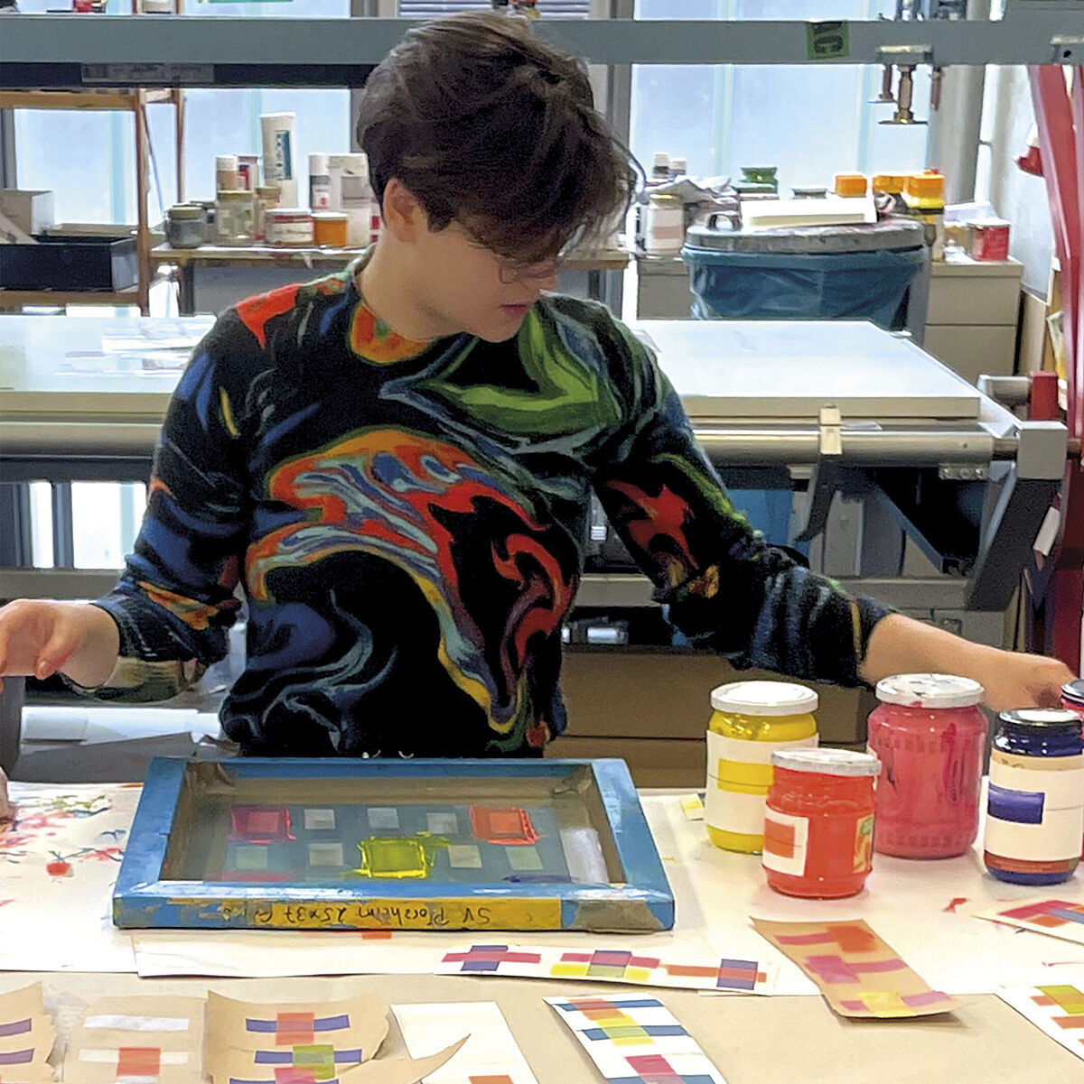 Eine Studentin, die einen butnen Pullover anhat, sitzt hinter einem großem Tisch in einem Atelier. Vor ihr liegen Papierstreifen mit verschiedenen bunten Mustern. Sie greift nach Farbtöpfen.