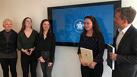 Es sind die drei Studentinnen, die Professorin und der Bürgermeister vor einem Bildschirm zu sehen, auf dem das Logo eingeblendet ist.