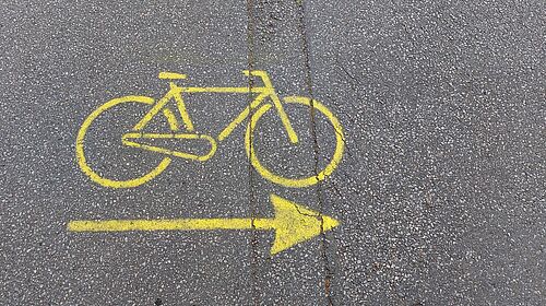 Es ist ein kleiner Straßenabschnitt von oben zu sehen, auf dessen Bitumenoberfläche sich eine Fahrradgrafik mit Pfeil befindet. Dises wurde mit gelber Farbe gezeichnet.