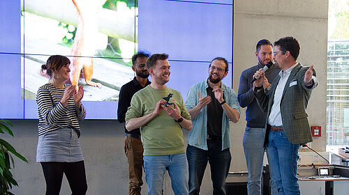 Rechts unten am Bildrand sieht man die klatschenden Hände des Publikums. Auch die sechs Personen auf der Bühne klatschen, während der Sieger einen Fischer-Pokal in den Händen hält.
