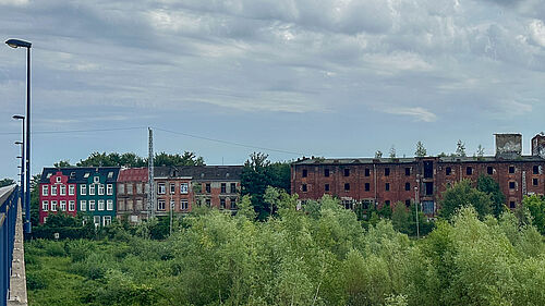 barrierefrei: Es ist ein verfallenes mehrstöckiges Backsteingebäude mit teilweise zerstörten Fenstern und angrenzenden bunten Wohnhäusern zu sehen - vor bewölktem Himmel und grüner Vegetation im Vordergrund.