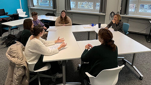 Es sind vier Studierende und zwei Dozentinnen  zu sehen, die an einem kreisförmig angeordneten Tisch im Seminarraum sitzensitzen