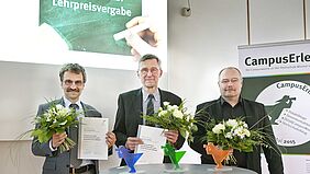 Die drei Professoren stehen mit Blumensträußen hinter einem Stehtisch, auf dem sich diedrei Lehrpreis (Fischerfiguren in Fakultätsfarbe) befinden.