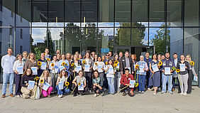 Eine Gruppe von Menschen steht oder hockt in mehreren Reihen vor einem modernen Glasgebäude. Viele halten Urkunden und Sonnenblumen in den Händen.