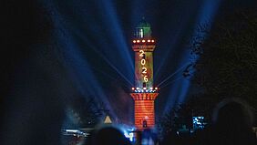 Der Warnemünder Leuchtturm ist rötlich angestrahlt. An ihm leuchten von oben nach untern die vier Ziffern 2, 0, 2 und 6 in Gelb.