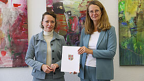 Zwei Frauen stehen vor bunten Gemälden. Eine hält eine Mappe mit dem Wappen M-Vs in der Hand.