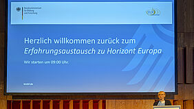 Eine Frau steht in der rechten unteren Ecke des Bildes an einem Pult. Hinter ihr ist ein Präsentationsbild mit dem Titel der Veranstaltung zu sehen.
